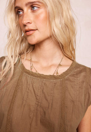 A close-up of a blonde woman wearing a brown textured top with lace trim and layered gold necklaces.